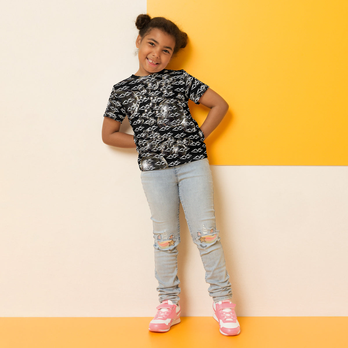Young girl wearing a black patterned shirt, light blue jeans, and pink sneakers against a yellow and beige background.